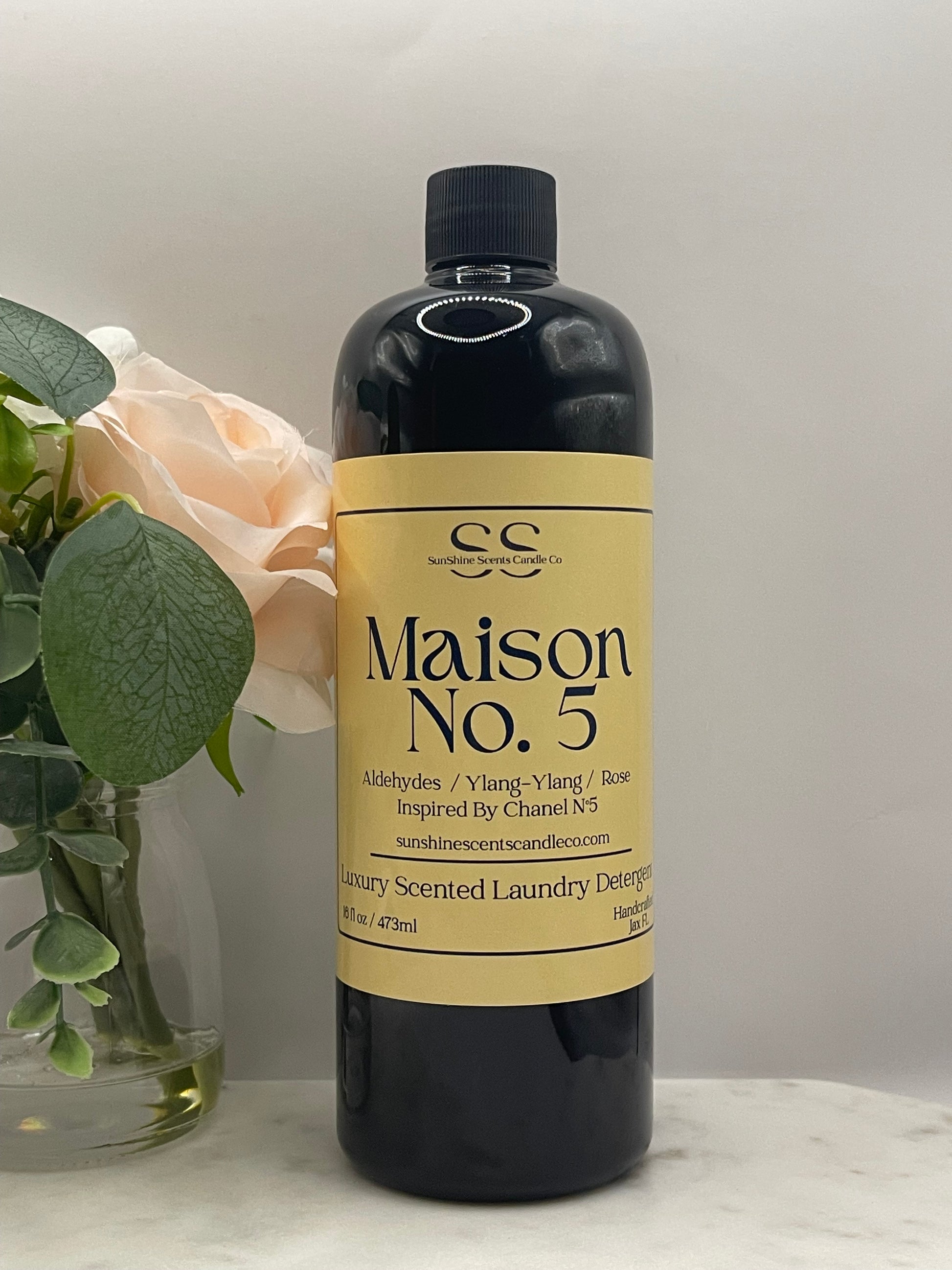 A bottle of Maison No. 5 Scented Laundry Purgative, displayed horizontally on a white marble surface with a pale peach rose and green leaves in the foreground.