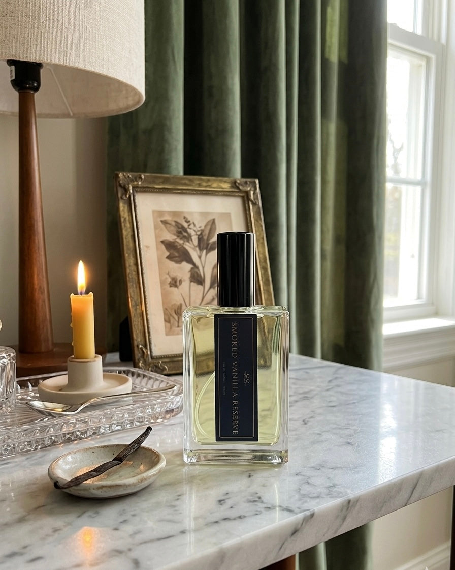 Smoked Vanilla Reserve Eau de Parfum 100ml bottle by SunShine Scents Candle Co. on a marble surface with a lit candle, vanilla bean, and a gold-framed botanical print." 