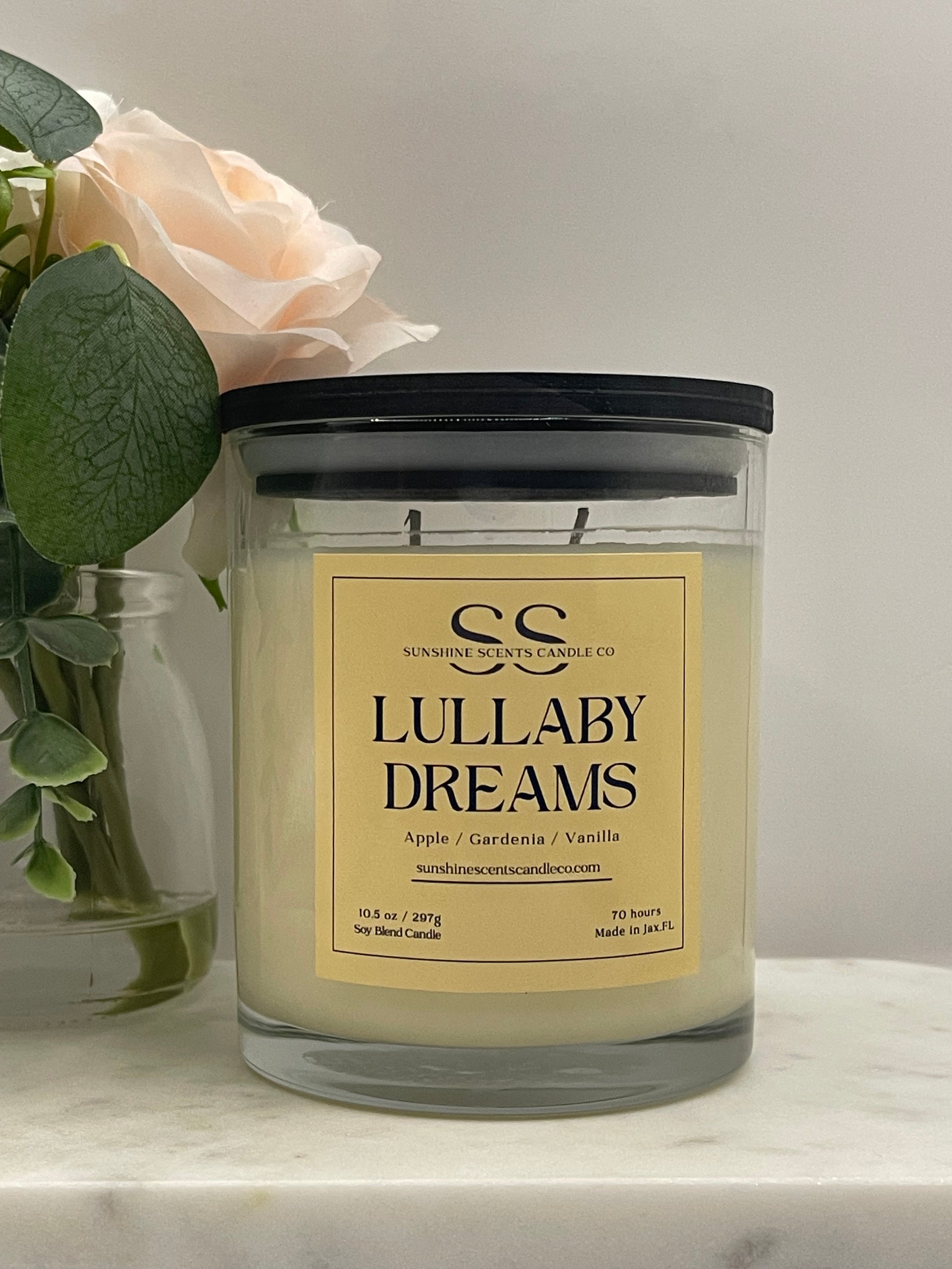 A glass jar candle with a black lid and a cream-colored label reading 'Lullaby Dreams' by Sweeteness Candle Co, styled with a pale peach rose and green leaves on a light marble surface.