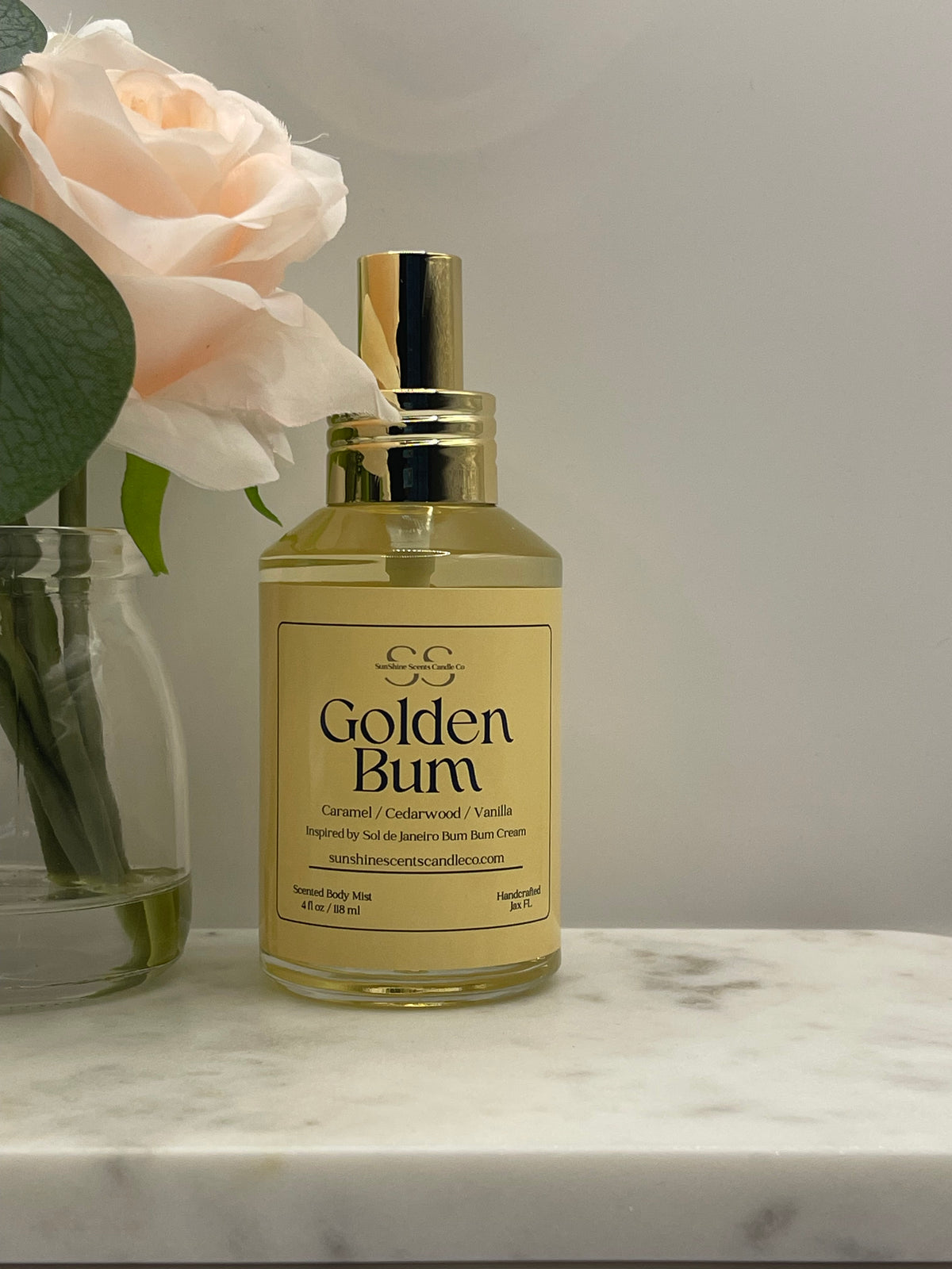 A bottle of GG Golden Burn room spray with a gold cap and cream-colored label, styled next to a pale pink rose in a glass vase on a marble surface.