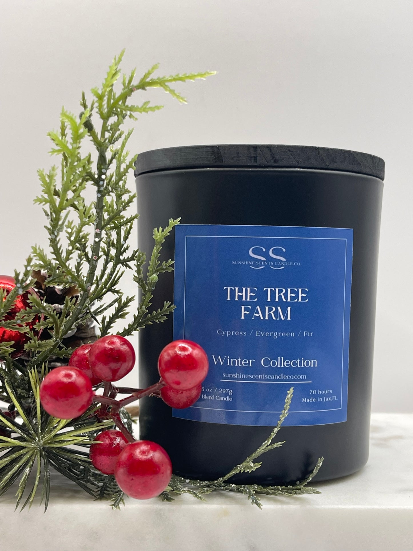 A black candle jar with a blue label reading 'The Tree Farm Winter Collection' and 'Cypress / Evergreen / Fir,' styled with evergreen branches and red berries on a light neutral background.