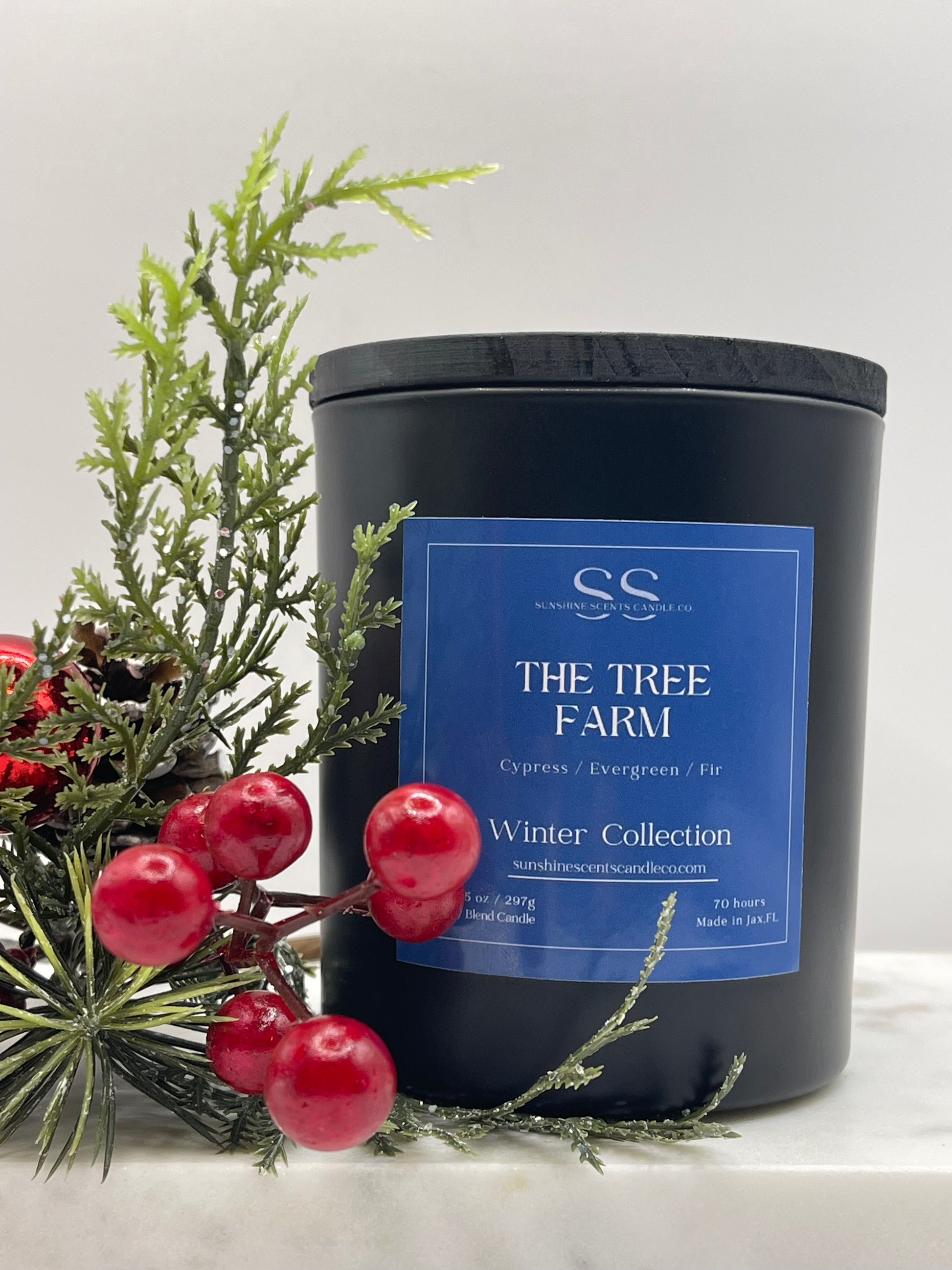 A black candle jar with a blue label reading 'The Tree Farm Winter Collection' and 'Cypress / Evergreen / Fir,' styled with evergreen branches and red berries on a light neutral background.