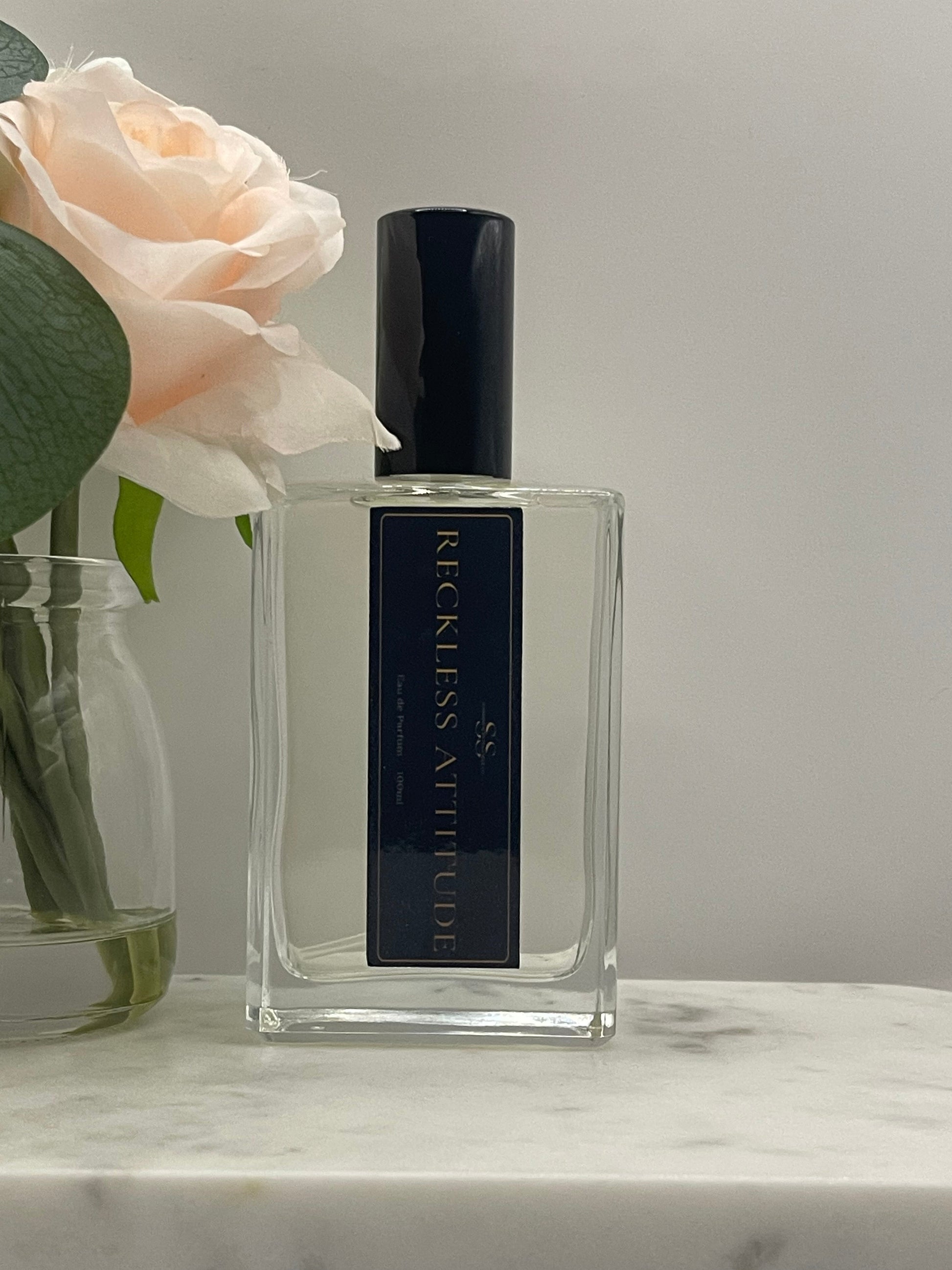 A clear rectangular glass perfume bottle with a black cap and a black label reading 'RECKLESS ATTITUDE' in gold lettering, styled with a pale peach rose and green leaves against a light gray background.