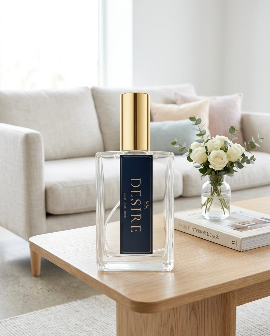 "Desire Eau de Parfum 100ml bottle by SunShine Scents Candle Co. with a gold cap on a wooden coffee table with white roses in a glass vase, an interior design book, and a soft grey sofa in the background." 