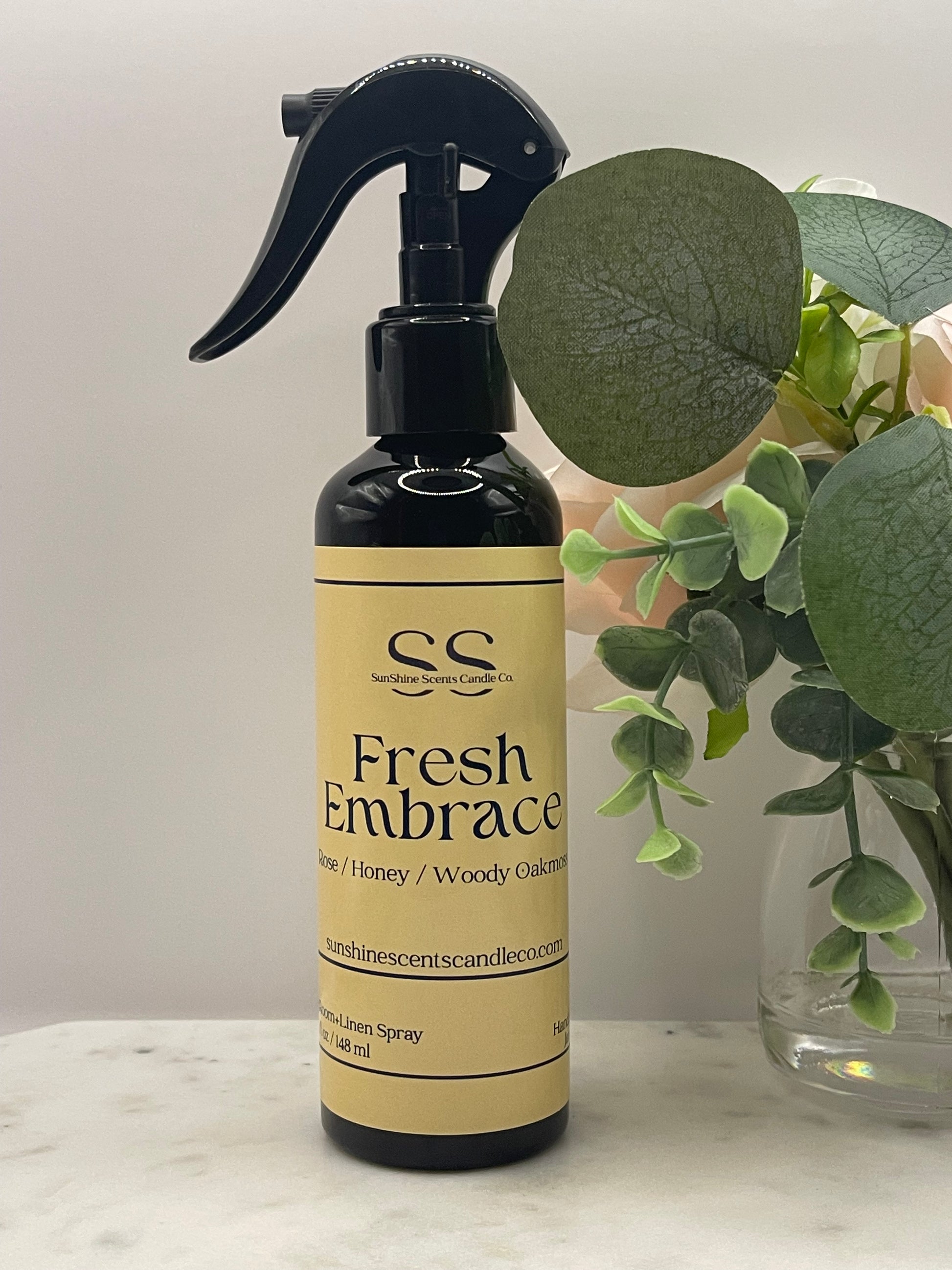 Fresh Embrace Room + Linen Spray by SunShine Scents Candle Co. — a black spray bottle with a dandelion-yellow label featuring Rose, Honey, and Woody Oakmoss scent notes, styled on a marble surface with eucalyptus and florals"