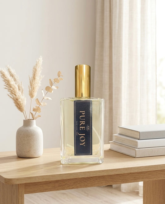 "Pure Joy Eau de Parfum 100ml bottle by SunShine Scents Candle Co. with a gold cap on a wooden shelf beside a ceramic vase with dried pampas grass and eucalyptus, with stacked white books and sheer curtains in the background." 