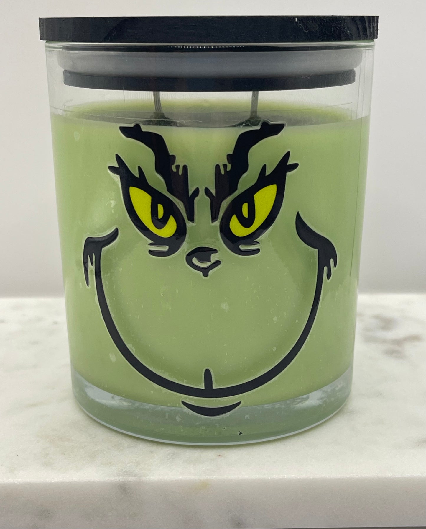 A green Grinch-themed candle in a clear glass jar with a black lid, featuring a printed face design with yellow eyes and black eyebrows and mouth details.