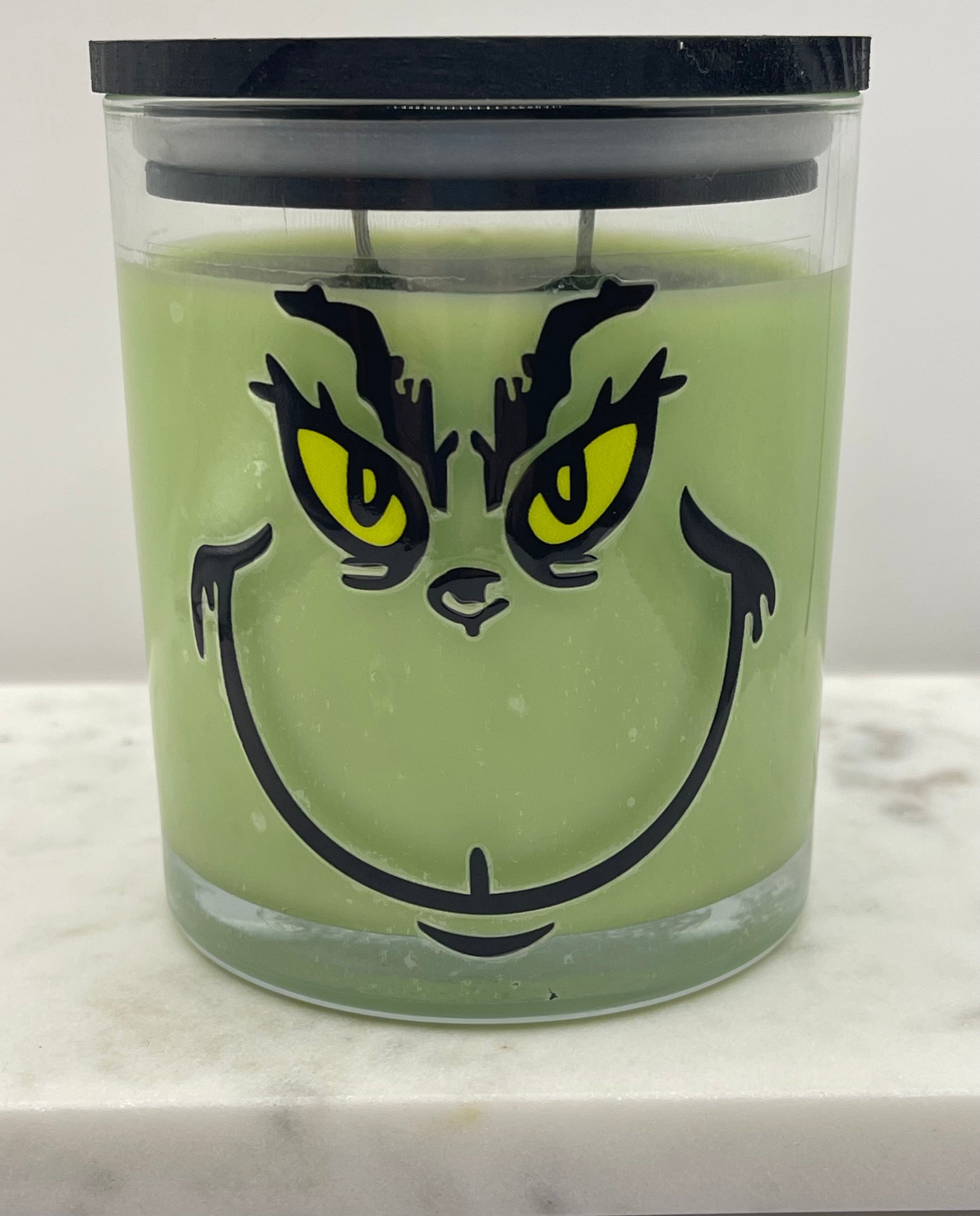 A green Grinch-themed candle in a clear glass jar with a black lid, featuring a printed face design with yellow eyes and black eyebrows and mouth details.