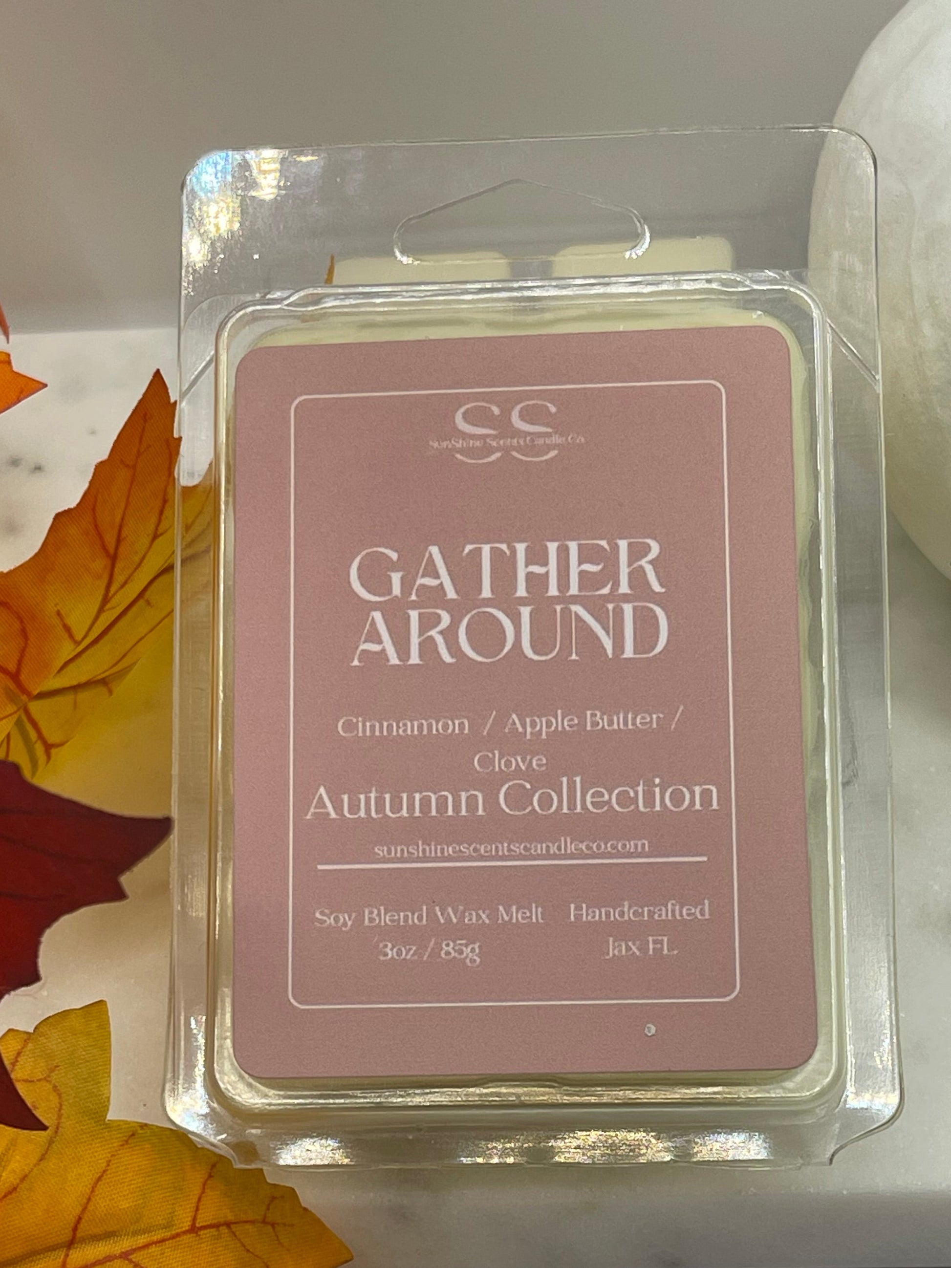 A wax melt package labeled 'Gather Around' by SS, placed on a white marble-like surface with autumn maple leaves around it.