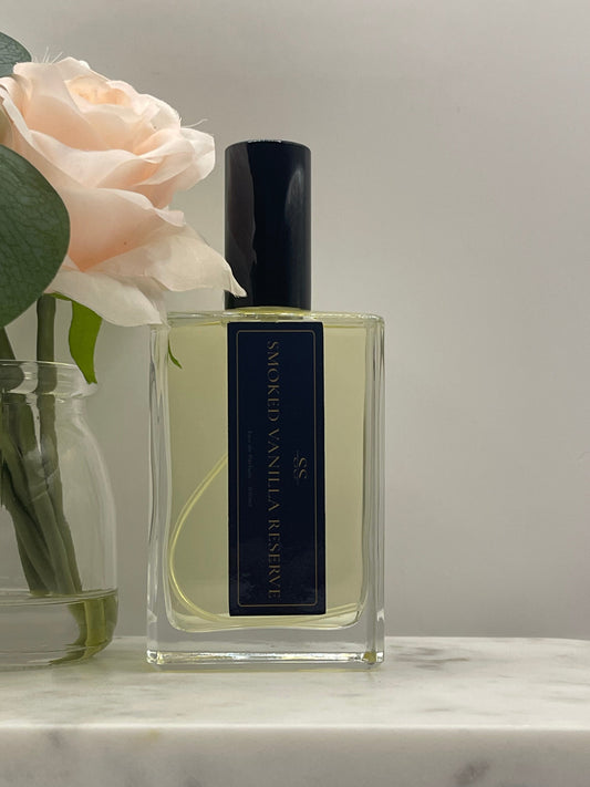 A rectangular glass perfume bottle labeled 'SMOKED VANILLA RESERVE' with a black cap, shown on a reflective surface with a pale peach rose and green leaves in the foreground.