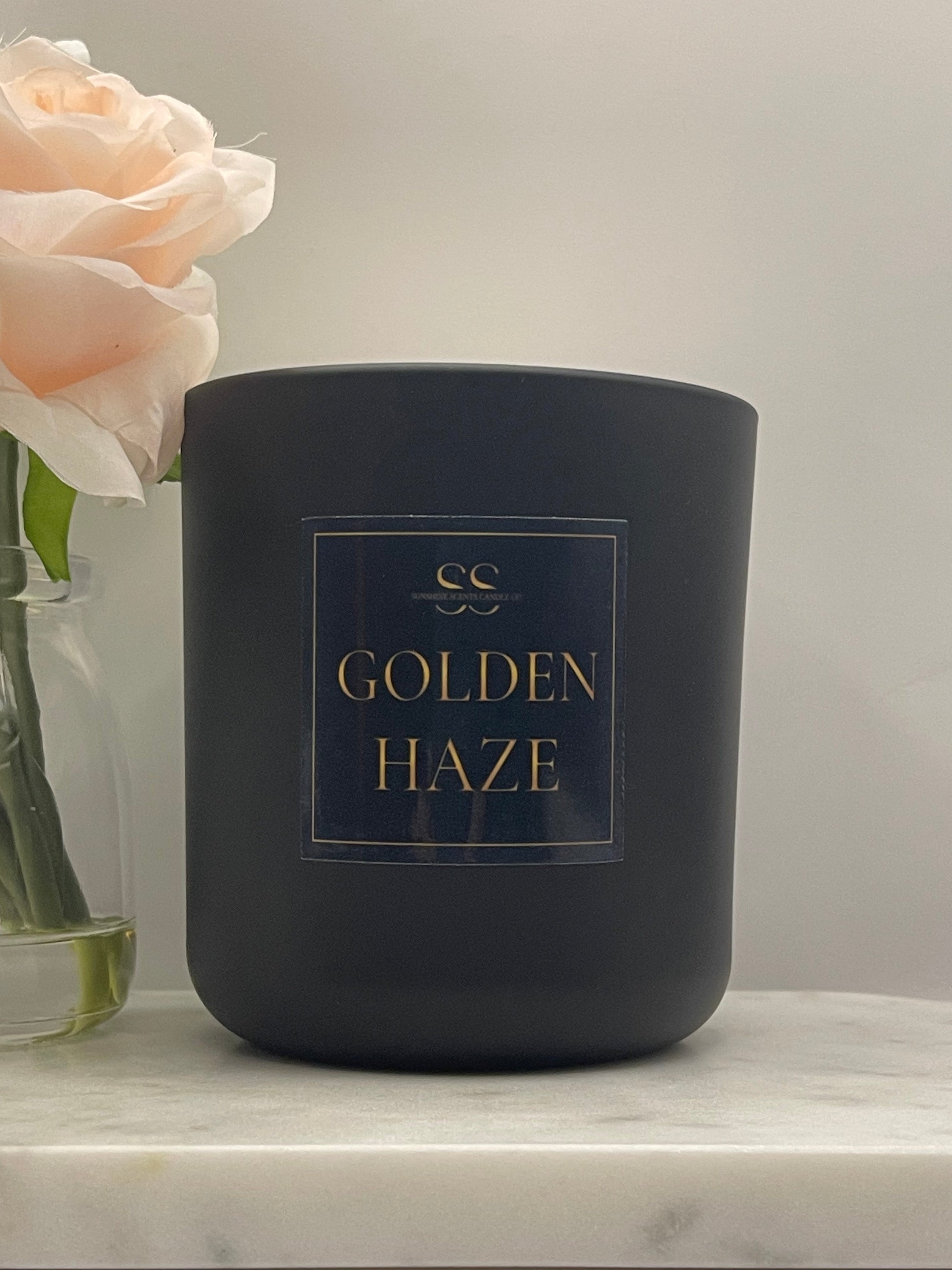 A matte black candle jar with a gold label reading 'Golden Haze' and a small CC logo, photographed on a light marble surface with pale pink flowers in the foreground.