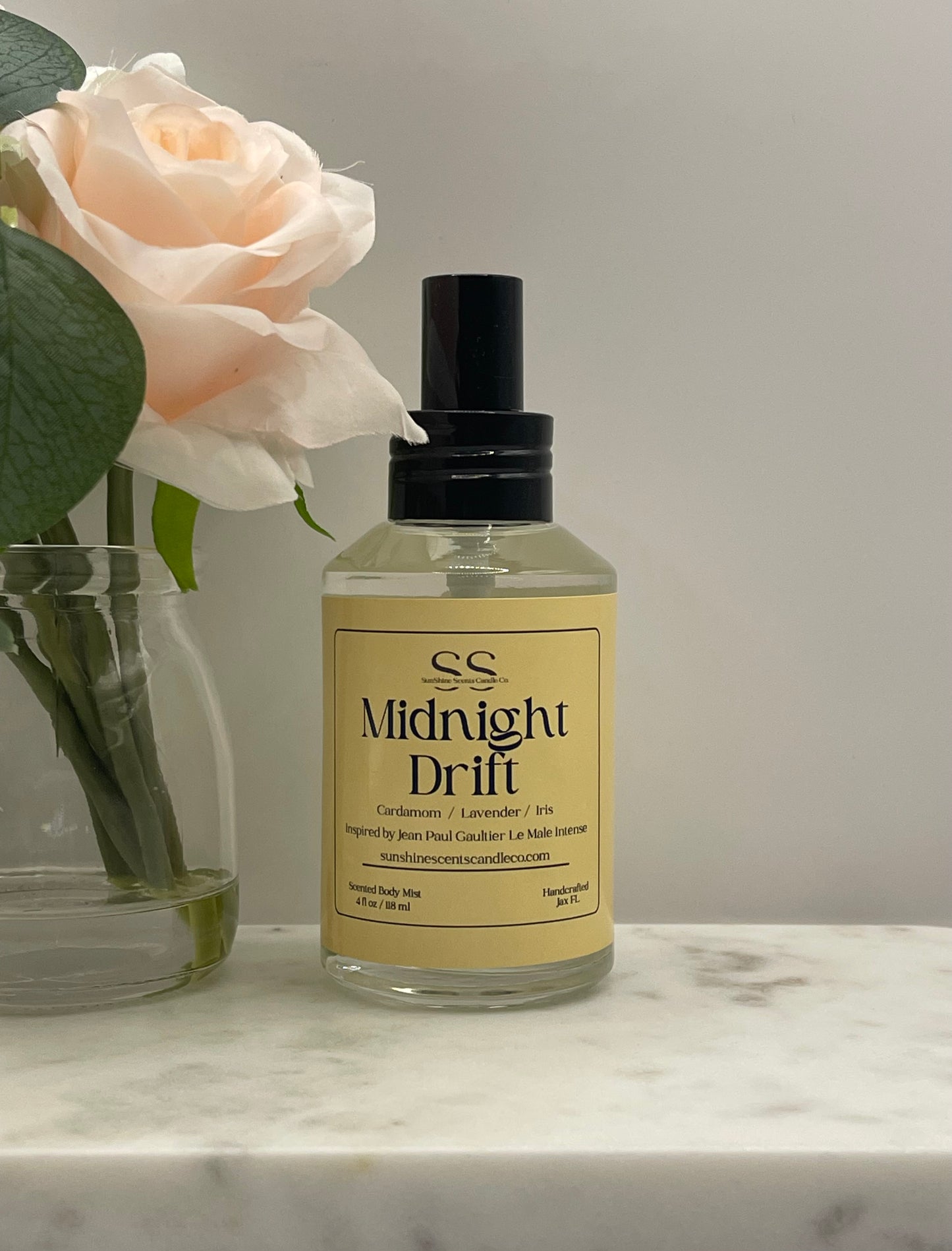 A bottle of 'Midnight Drift' room spray by SS, featuring a pale yellow label and black spray cap, set on a white marble surface next to a glass vase with a pale peach rose.