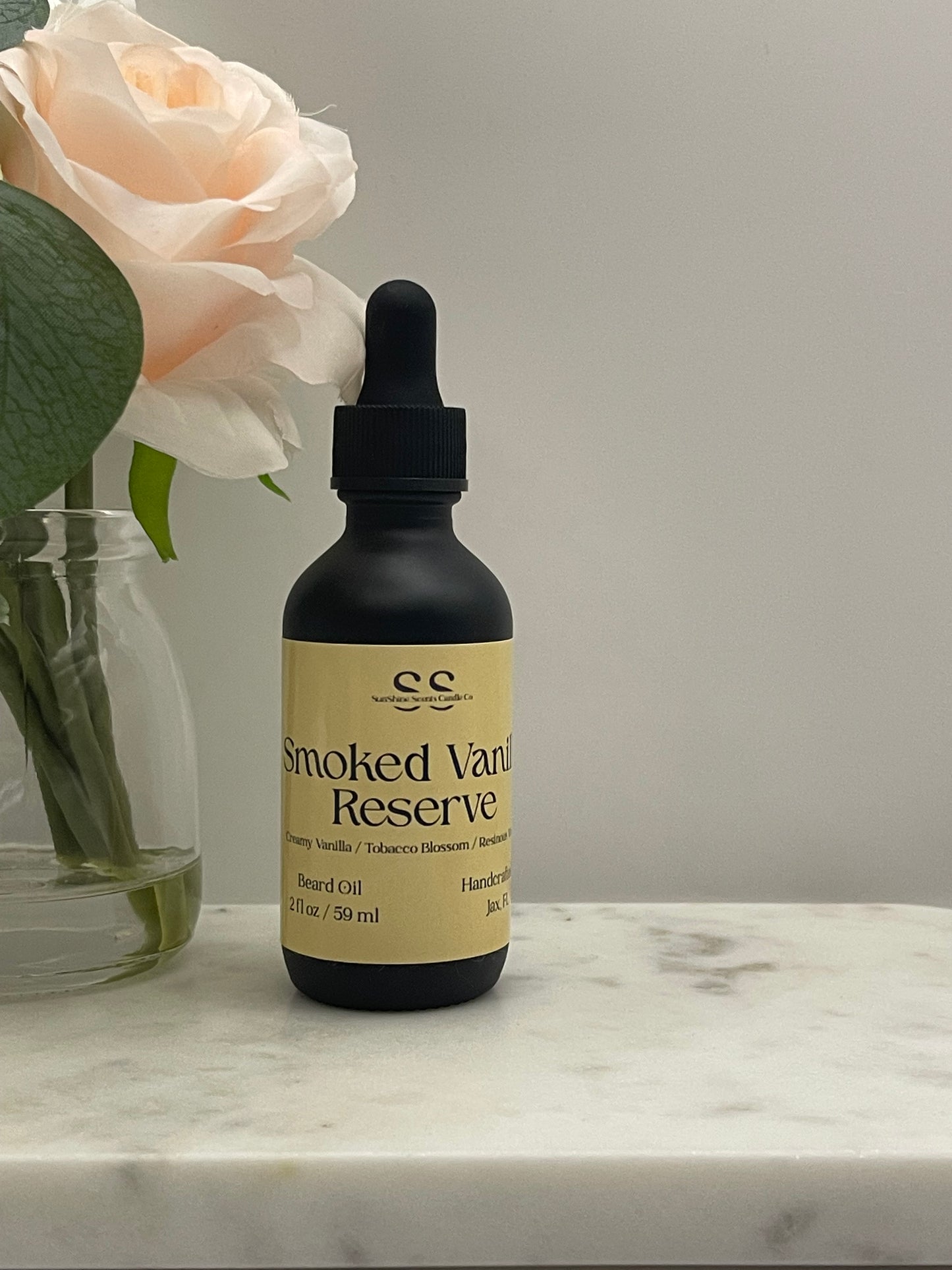 A dark amber glass dropper bottle with a beige label reading 'Smoked Vanilla Reserve' by the brand 'sc', resting on a white marble surface next to a pale peach rose in a glass vase.