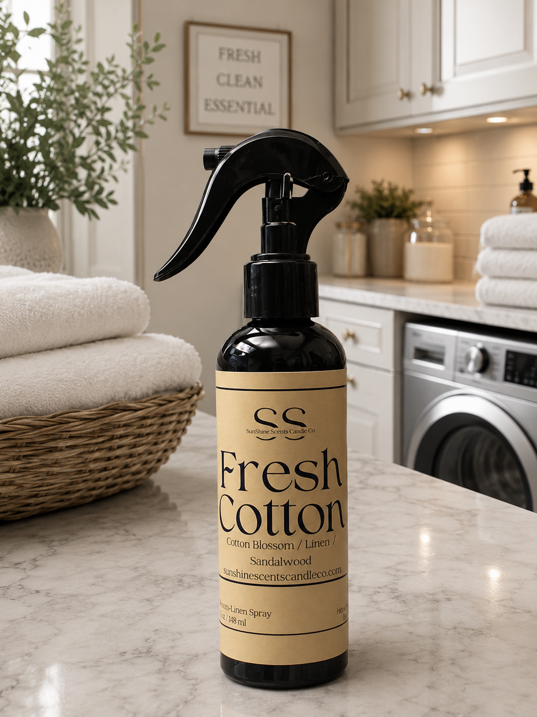A dark amber spray bottle labeled 'Fresh Cotton' with a beige label and black trigger sprayer, placed on a marble countertop in a bright laundry room with folded towels and a washing machine in the background.