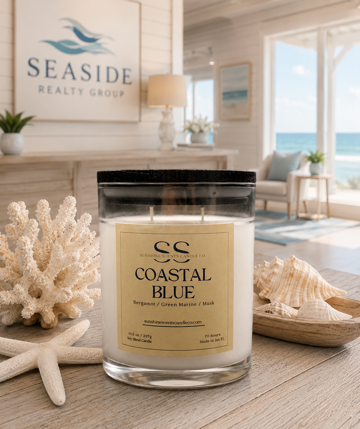 A jar candle labeled 'Coastal Blue' by Seaside Realty Group is displayed on a wooden table in a bright, beach-themed room. The candle is styled with seashells, coral, and a starfish, with a blurred background showing an ocean view.