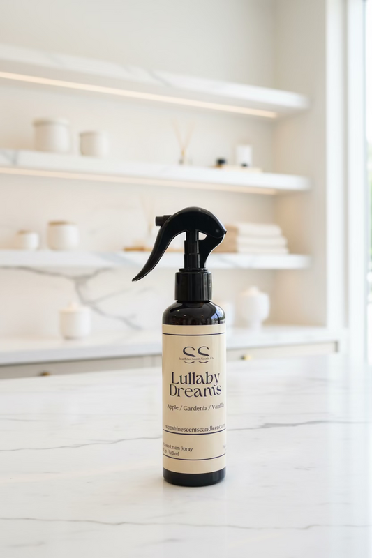 A dark amber spray bottle with a black trigger sprayer and a cream label reading 'SS Lullaby Dreams' stands on a white marble countertop in a softly blurred, warm-toned interior with shelves in the background.