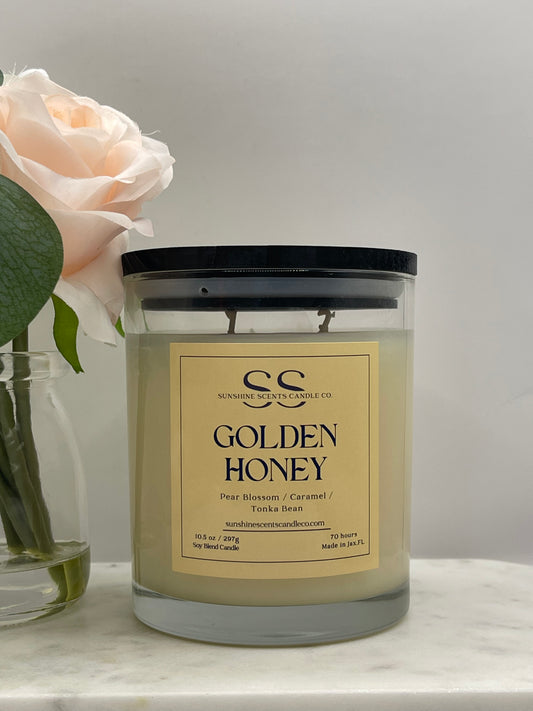 Golden Honey 10.5 oz soy blend candle by SunShine Scents Candle Co. in a clear glass jar with a black lid, featuring a gold label with notes of Pear Blossom, Caramel, and Tonka Bean, styled on a marble surface with a blush pink rose